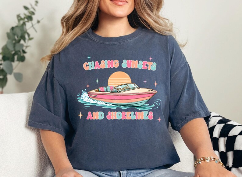 Pu&ograve; includere: T-shirt blu navy con grafica colorata di una barca sull'acqua, un tramonto e la scritta "Chasing Sunsets and Shorelines". Il design utilizza colori pastello e stelle.
