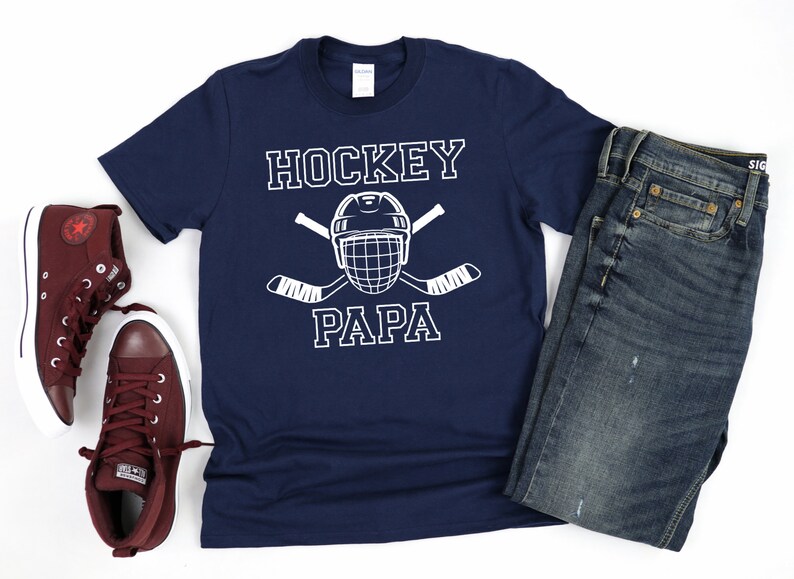 May include: Navy blue t-shirt with the text "HOCKEY PAPA" and a hockey helmet and crossed hockey sticks graphic. The shirt is paired with blue jeans and burgundy high-top sneakers.
