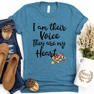 K&ouml;nnte beinhalten: Ein t&uuml;rkisfarbenes T-Shirt mit dem Text "I am their Voice They are my Heart" und einem Puzzle-Herz-Design. Das Shirt liegt auf einer wei&szlig;en Oberfl&auml;che mit Jeans, Schuhen und einer Blumenvase.