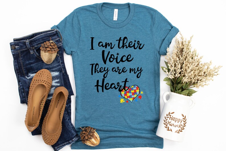 K&ouml;nnte beinhalten: Ein t&uuml;rkisfarbenes T-Shirt mit dem Text "I am their Voice They are my Heart" und einem Puzzle-Herz-Design. Das Shirt liegt auf einer wei&szlig;en Oberfl&auml;che mit Jeans, Schuhen und einer Blumenvase.