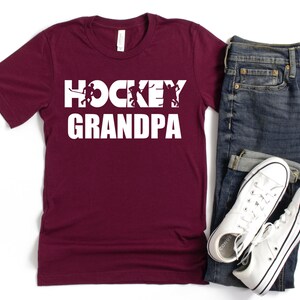 May include: Burgundy t-shirt with white text that reads "Hockey Grandpa" and features a silhouette of hockey players.