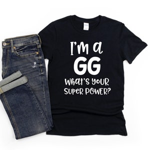 May include: Black t-shirt with white text that reads "I'm a GG What's your super power?" The shirt is laying on a white surface next to a pair of blue jeans.