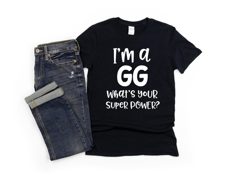 May include: Black t-shirt with white text that reads "I'm a GG What's your super power?" The shirt is laying on a white surface next to a pair of blue jeans.