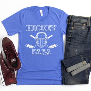 May include: A blue t-shirt with the words "HOCKEY PAPA" in white, along with a hockey helmet and crossed hockey sticks. The shirt is paired with dark wash jeans and burgundy high-top sneakers.