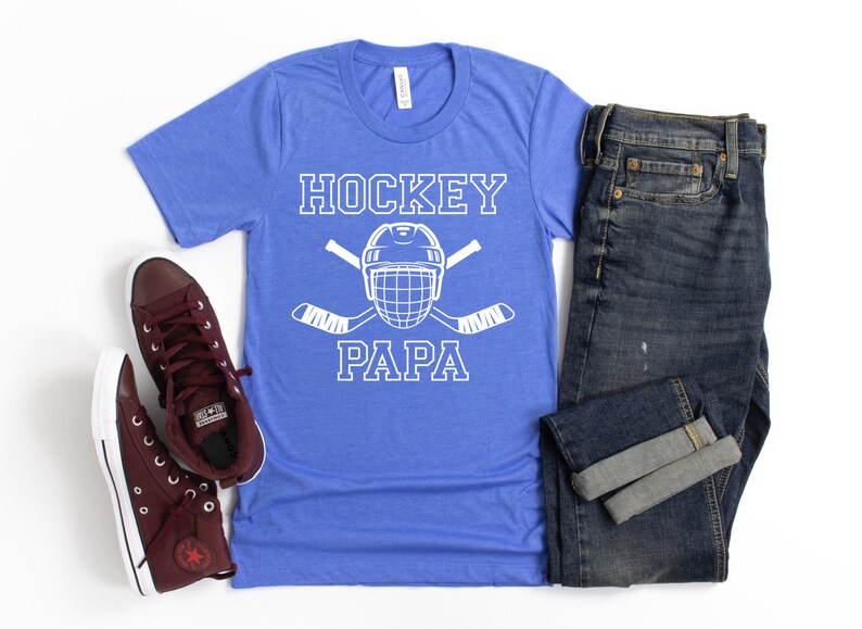 May include: A blue t-shirt with the words "HOCKEY PAPA" in white, along with a hockey helmet and crossed hockey sticks. The shirt is paired with dark wash jeans and burgundy high-top sneakers.