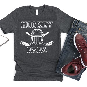 May include: Dark gray t-shirt with the words "HOCKEY PAPA" in white block letters, along with a hockey helmet and crossed hockey sticks. The shirt is paired with blue jeans and maroon sneakers.