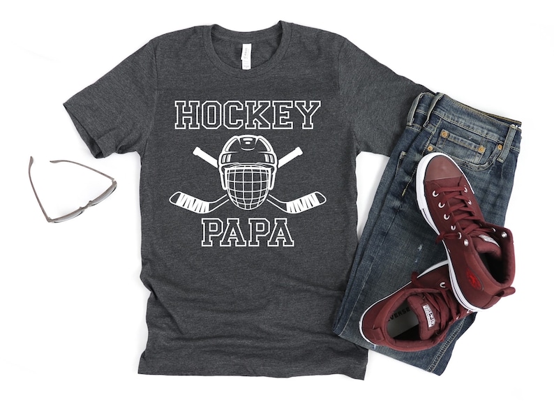 May include: Dark gray t-shirt with the words "HOCKEY PAPA" in white block letters, along with a hockey helmet and crossed hockey sticks. The shirt is paired with blue jeans and maroon sneakers.