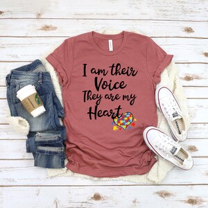 K&ouml;nnte beinhalten: Ein maronfarbenes T-Shirt mit dem Text "I am their Voice They are my Heart" und einem Herz mit einem Puzzle-St&uuml;ck-Design. Das Shirt liegt auf einer wei&szlig;en Oberfl&auml;che mit einem Paar wei&szlig;en Sneakers und einer blauen Jeans.