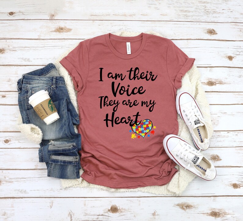 K&ouml;nnte beinhalten: Ein maronfarbenes T-Shirt mit dem Text "I am their Voice They are my Heart" und einem Herz mit einem Puzzle-St&uuml;ck-Design. Das Shirt liegt auf einer wei&szlig;en Oberfl&auml;che mit einem Paar wei&szlig;en Sneakers und einer blauen Jeans.