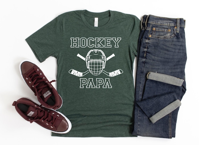 May include: Green t-shirt with the text "HOCKEY PAPA" in white, featuring a hockey helmet and crossed hockey sticks. The shirt is paired with blue jeans and maroon sneakers.