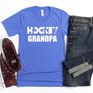 May include: A royal blue t-shirt with white text that reads "HOCKEY GRANDPA" with a graphic of hockey players. The shirt is displayed with a pair of maroon high-top sneakers and a pair of blue jeans.