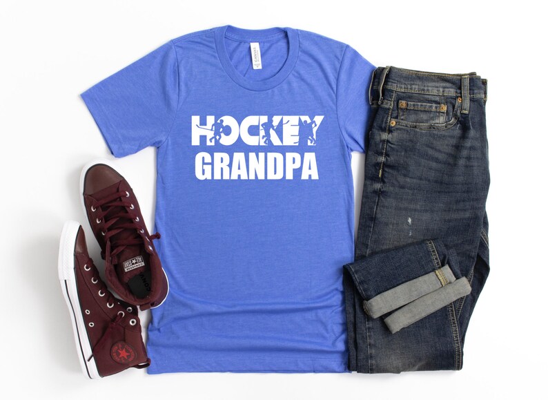 May include: A royal blue t-shirt with white text that reads "HOCKEY GRANDPA" with a graphic of hockey players. The shirt is displayed with a pair of maroon high-top sneakers and a pair of blue jeans.
