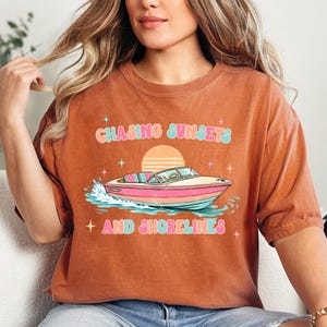 Pu&ograve; includere: T-shirt color ruggine con grafica di una barca sull'acqua, un tramonto e la scritta "Chasing Sunsets and Shorelines" in colori pastello. La maglietta ha una vestibilit&agrave; ampia.