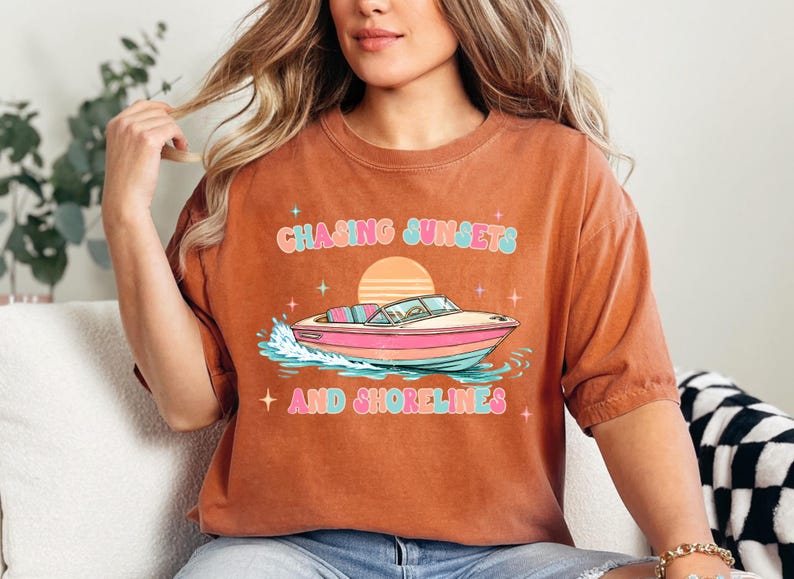Pu&ograve; includere: T-shirt color ruggine con grafica di una barca sull'acqua, un tramonto e la scritta "Chasing Sunsets and Shorelines" in colori pastello. La maglietta ha una vestibilit&agrave; ampia.