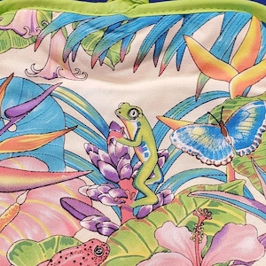 May include: A green frog sits on a purple flower with a blue butterfly on a green leaf. A pink frog sits on a green leaf with a pink hibiscus flower. The background is a white fabric with a tropical leaf pattern. The text "©agiftcorp" is in the bottom left corner.