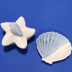 May include: Two ceramic trinket dishes, one shaped like a starfish and the other like a seashell. Both are white with blue accents.