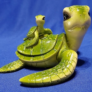 Puede incluir: Una figurita de tortuga de cerámica verde con una figurita de tortuga verde más pequeña montada en su espalda. Ambas tortugas tienen detalles realistas y ojos pintados.
