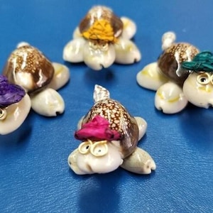 May include: Five whimsical turtle figurines with shell bodies and hats. The turtles have white bodies, brown shells, and are adorned with small hats in various colors, including purple, yellow, and pink. They have gold-colored wire-rimmed glasses. The figurines are on a blue surface.