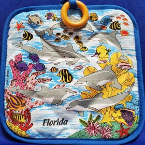 Peut inclure: Un dessous de plat bleu et blanc avec une scène sous-marine colorée représentant des dauphins, des coraux et des poissons. Le dessous de plat est doté d'un anneau en bois fixé en haut et du texte "Florida" imprimé en bas.