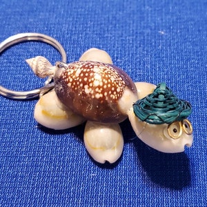 May include: A seashell turtle keyring with a green woven hat and gold-rimmed glasses. The turtle is made of various seashells and has a silver key ring.