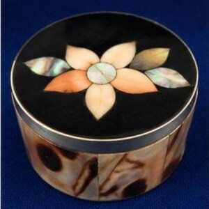 May include: A round box with a black lid featuring an inlaid flower design made of iridescent shell petals in shades of cream and peach. The sides of the box are covered in a mosaic of polished, mottled brown and tan shell segments.