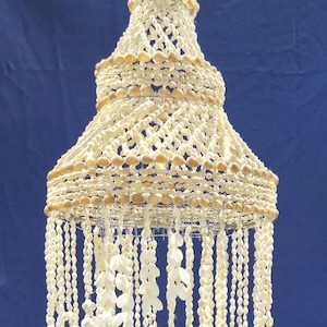 May include: A tiered hanging light fixture constructed from a wire frame covered in small, off-white seashells. The upper section features a woven shell pattern, while the lower portion consists of numerous long, dangling strands of white shells.