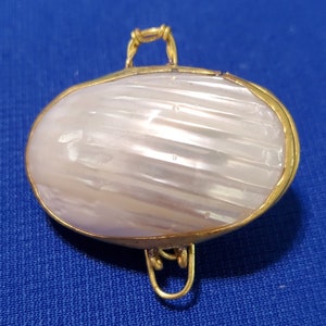 Pode incluir: Uma pequena caixa de lembranças oval de madrepérola branca com uma moldura de metal dourado e uma tampa com dobradiças. A caixa tem uma alça de arame decorativa.