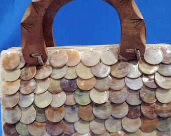 May include: A rectangular handbag featuring a beige fabric base covered in overlapping circular, iridescent shell discs in shades of brown, tan, and cream. The bag includes a semi-circular, carved dark brown wooden handle with etched details.