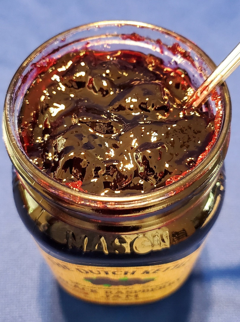 May include: A glass jar filled with dark red raspberry jam. The jar has a gold lid and a label that reads "The Dutch Kettle Black Raspberry Jam".