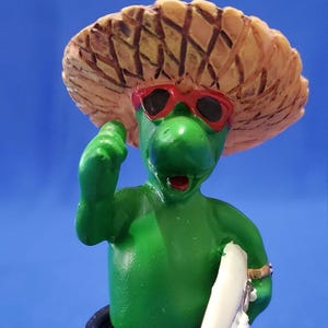 May include: A green turtle figurine wearing red sunglasses and a straw hat, holding a white surfboard. The turtle is smiling and has its arm raised in a thumbs up gesture.