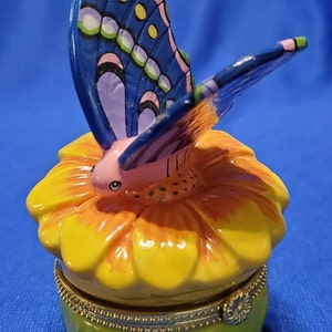 May include: A ceramic trinket box shaped like a yellow flower with a blue butterfly perched on top. The butterfly has pink, green, and yellow accents.
