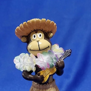 May include: A ceramic monkey figurine wearing a straw hat, lei, and grass skirt, playing a yellow ukulele. The monkey has a tan face, brown arms, and a brown body. The background is a solid blue.