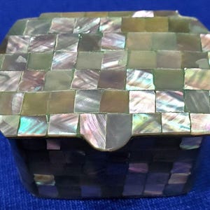 May include: A small rectangular box featuring a lid and body covered in a mosaic of iridescent mother-of-pearl tiles. The tiles display a variety of green, brown, and pearlescent hues with a reflective finish, arranged in a grid pattern.