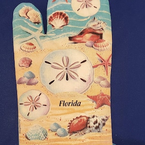 Puede incluir: Un guante de horno azul y blanco con una escena de playa con conchas marinas, estrellas de mar y dólares de arena. El texto "Florida" está impreso en el guante.
