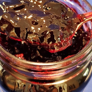 May include: A glass jar filled with dark purple blackberry jam. The jam is thick and glossy, and a spoonful is being lifted out of the jar. The jar has a label that says "The Dutch Kettle".