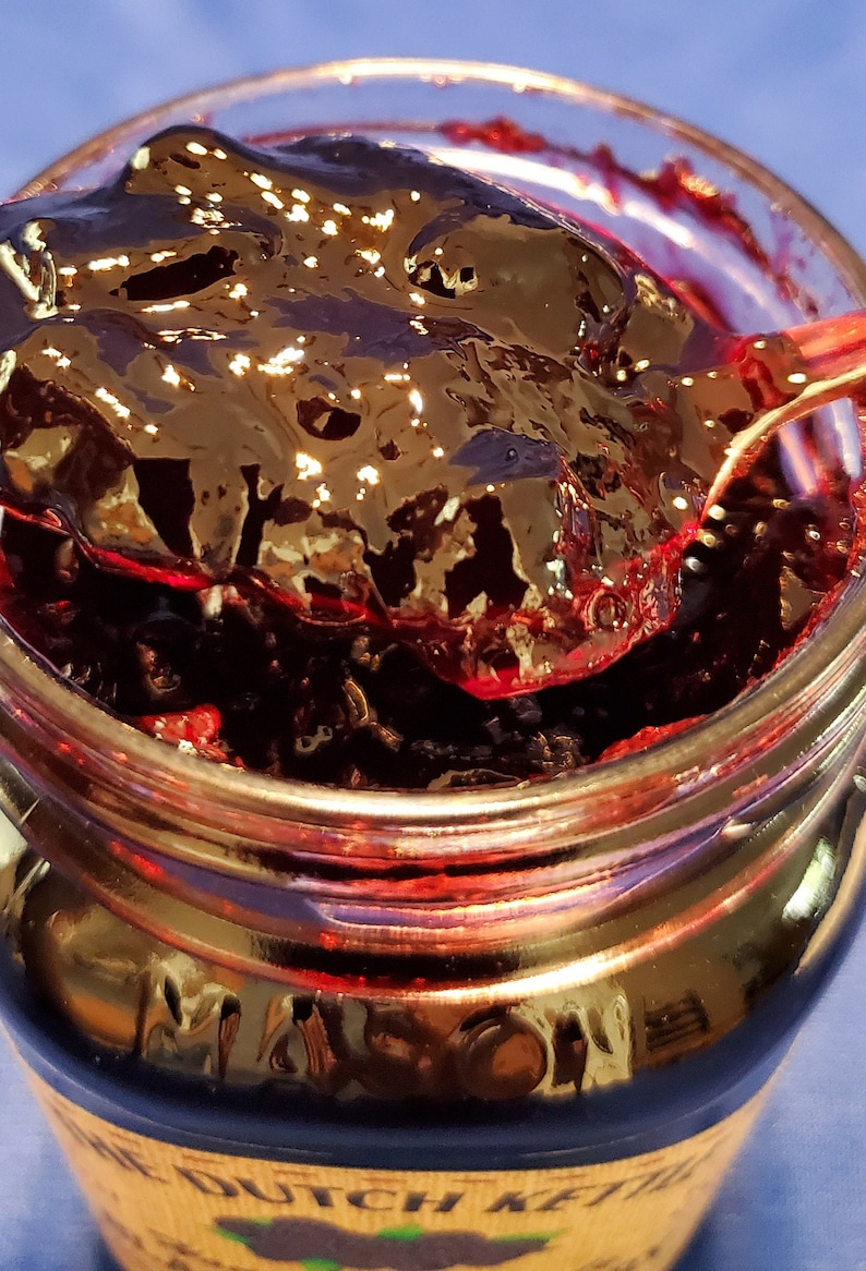 May include: A glass jar filled with dark purple blackberry jam. The jam is thick and glossy, and a spoonful is being lifted out of the jar. The jar has a label that says "The Dutch Kettle".