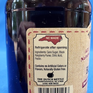 May include: A glass jar with a black label that reads "NC MADE" and "THE DUTCH KETTLE" with a phone number and address. The label also says "Refrigerate after opening" and lists the ingredients: Cane Sugar, Black Raspberry Puree, Citric Acid, Pectin. The label also says "Contains no Artificial Colors or Flavors, Naturally Gluten Free". The jar has a barcode and the number 8 91841 00125 3.