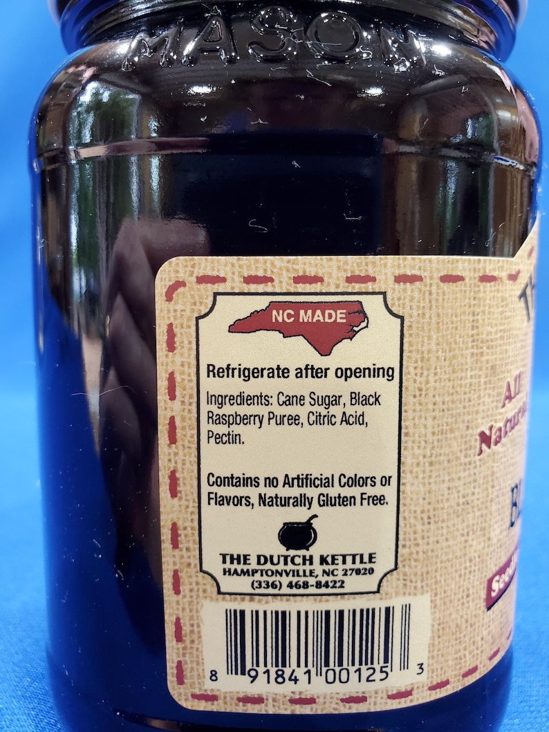 May include: A glass jar with a black label that reads "NC MADE" and "THE DUTCH KETTLE" with a phone number and address. The label also says "Refrigerate after opening" and lists the ingredients: Cane Sugar, Black Raspberry Puree, Citric Acid, Pectin. The label also says "Contains no Artificial Colors or Flavors, Naturally Gluten Free". The jar has a barcode and the number 8 91841 00125 3.