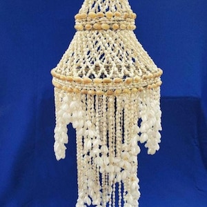Pode incluir: Um abajur suspenso construído com inúmeras pequenas conchas brancas dispostas num padrão de macramé em camadas. A estrutura apresenta uma parte superior cónica e uma larga faixa central circular, com longos fios de conchas em cascata.