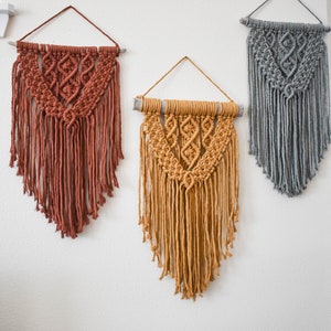 May include: Three macrame wall hangings with wooden dowels. The hangings are in shades of rust, mustard yellow, and gray.