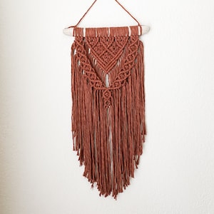 May include: A terracotta-colored macrame wall hanging. The handmade piece features intricate knotting patterns, a triangular design, and long, fringed tassels. It is suspended from a natural wooden dowel and a matching cord for hanging.