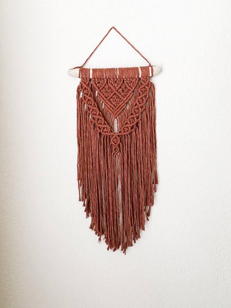 May include: A terracotta-colored macrame wall hanging. The handmade piece features intricate knotting patterns, a triangular design, and long, fringed tassels. It is suspended from a natural wooden dowel and a matching cord for hanging.