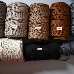 May include: A collection of spools of macrame cord in various colors, including storm gray, sand, mocha, maple, light gray, natural, black, and dark gray. Each spool is labeled with its color name.