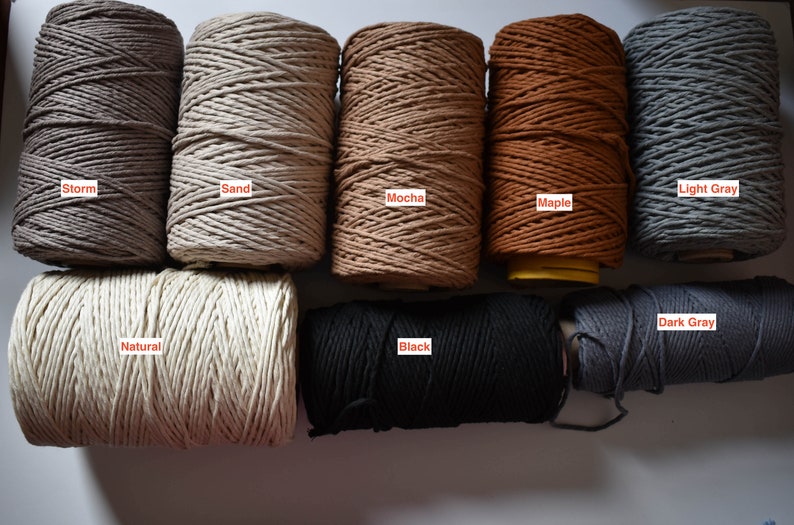 May include: A collection of spools of macrame cord in various colors, including storm gray, sand, mocha, maple, light gray, natural, black, and dark gray. Each spool is labeled with its color name.