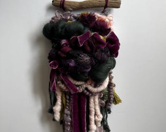 May include: A handcrafted fiber art wall hanging. The piece features a variety of textures and colors, including deep greens, purples, and pinks. It is suspended from a natural wooden branch with a dark cord.