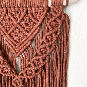 May include: A close-up of a handmade macrame wall hanging. The wall decor features a geometric design in a warm terracotta color. The macrame is attached to a natural wooden dowel, creating a bohemian style.