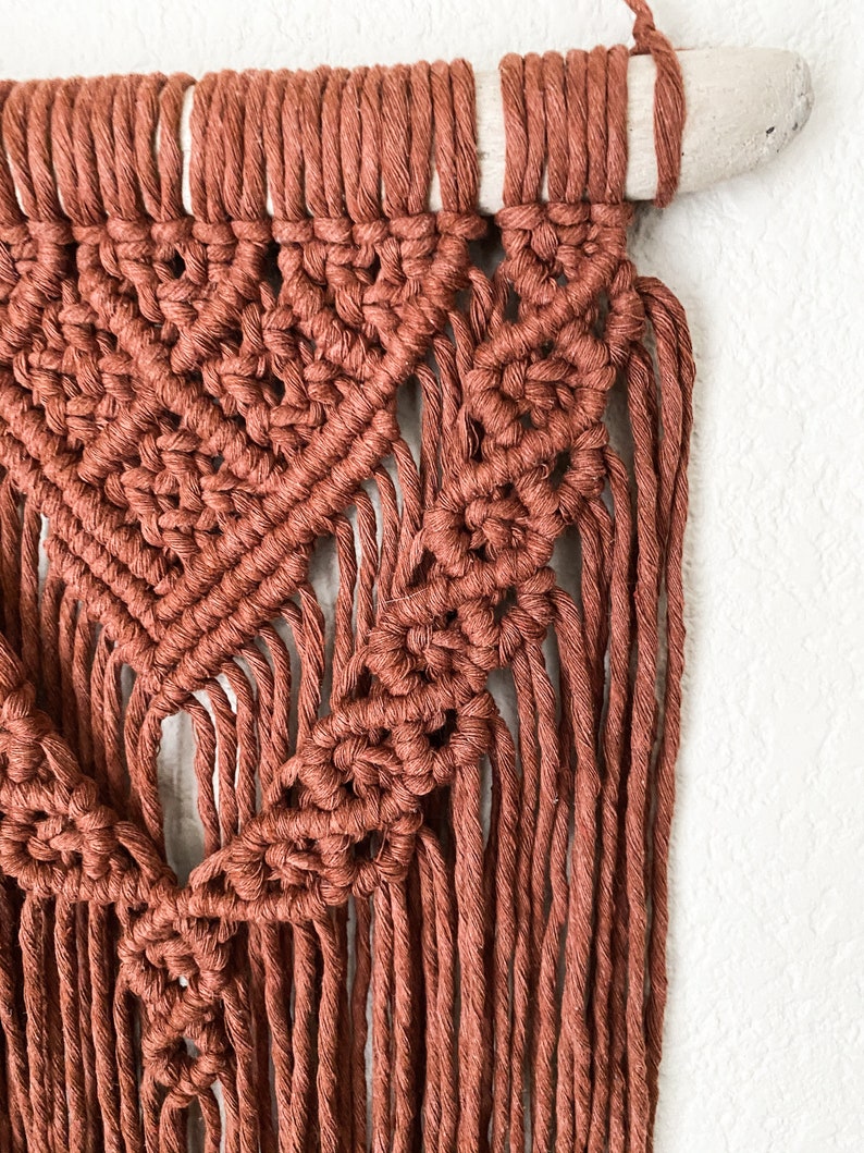 May include: A close-up of a handmade macrame wall hanging. The wall decor features a geometric design in a warm terracotta color. The macrame is attached to a natural wooden dowel, creating a bohemian style.