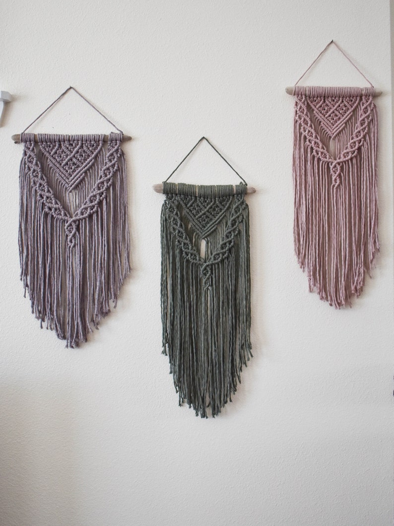 May include: Three macrame wall hangings in shades of gray and mauve. Each piece features a geometric design and fringe. They are suspended from wooden dowels with a triangular hanging cord, adding a touch of bohemian decor.