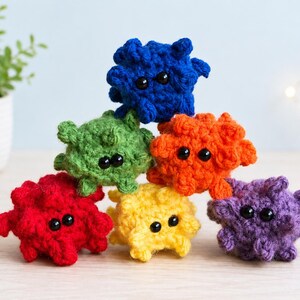 May include: A stack of six small, crocheted plush toys in vibrant colors including blue, green, orange, red, yellow, and purple. Each round, textured figure features two small, black, glossy plastic eyes and multiple protruding, nub-like appendages.