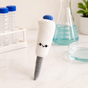 May include: A crochet pipette toy featuring a white body, a grey tip, and a blue top. The white section has two small black bead eyes and a stitched black mouth with pink blush marks. The toy is crafted with yarn in a soft, textured stitch pattern.
